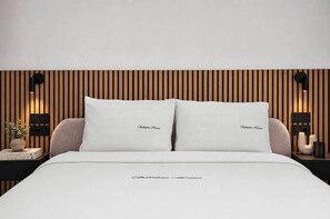 A modern bedroom with a wooden slat wall, two white pillows with 'Etrattore Homes' embroidered, a bedside table with a plant, books, and a black lamp. - NEW: Casa Serenia - Residence Corte Carden 2 (Bardolino)