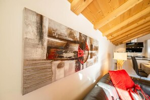 A modern living room with a large abstract painting on the wall, a leather sofa with red cushions, and a wooden ceiling. - Victoria C - InspiringPlaces (Zermatt)
