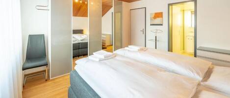Una habitación de hotel moderna con techo de madera, cama con ropa blanca, una silla y un baño a la vista.