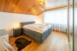 A bedroom with a wooden ceiling, a bed with white bedding, a nightstand, a fur rug, and a window with sheer curtains. - Victoria C - InspiringPlaces (Zermatt)