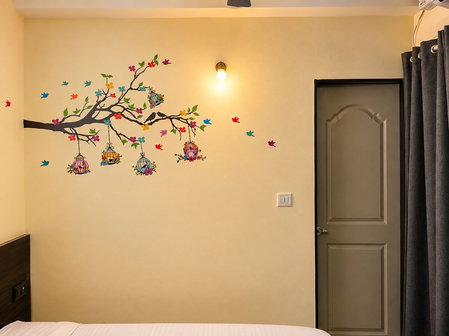 Una habitación con un adorno en la pared de un árbol con aves y flores de colores, una lámpara, una puerta con cortinas y una cama con ropa blanca.