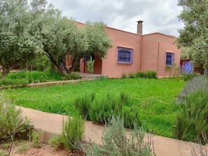 A building with a red exterior, a garden with green grass and trees, and a clear sky. - Villa Tissi house (Marrakech)