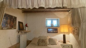 A bedroom with a bed, a nightstand with a lamp, a small window with a view, and a wall-mounted shelf with a vase and picture.