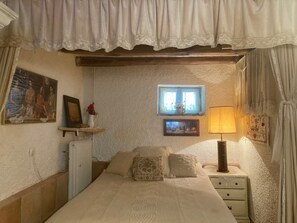 A bedroom with a bed, a nightstand with a lamp, a small window with a view, and a wall-mounted shelf with a vase and picture.