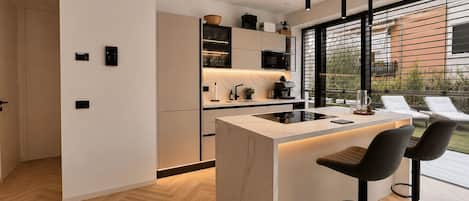 A modern kitchen with a central island, black bar stools, and a view of the outdoors through large windows.