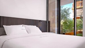 Une chambre d’hôtel moderne avec un grand lit, une tête de lit en bois et une vue sur la ville grâce à de grandes fenêtres du sol au plafond.