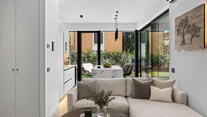 Un salon moderne avec un canapé, une table basse et un vase agrémenté de plantes vertes. On aperçoit également un coin repas avec une table et des chaises, visibles à travers une porte vitrée.