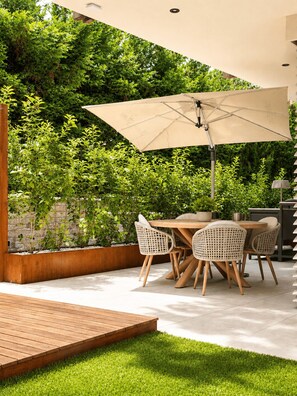 Eine Terrasse im Freien mit einem Holztisch und -stühlen, einem großen Sonnenschirm und üppiger Bepflanzung im Hintergrund.