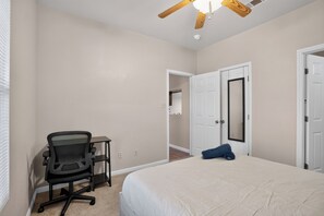 Ein Schlafzimmer mit Bett, Schreibtisch mit Stuhl, Deckenventilator und einer Tür zu einem anderen Raum.