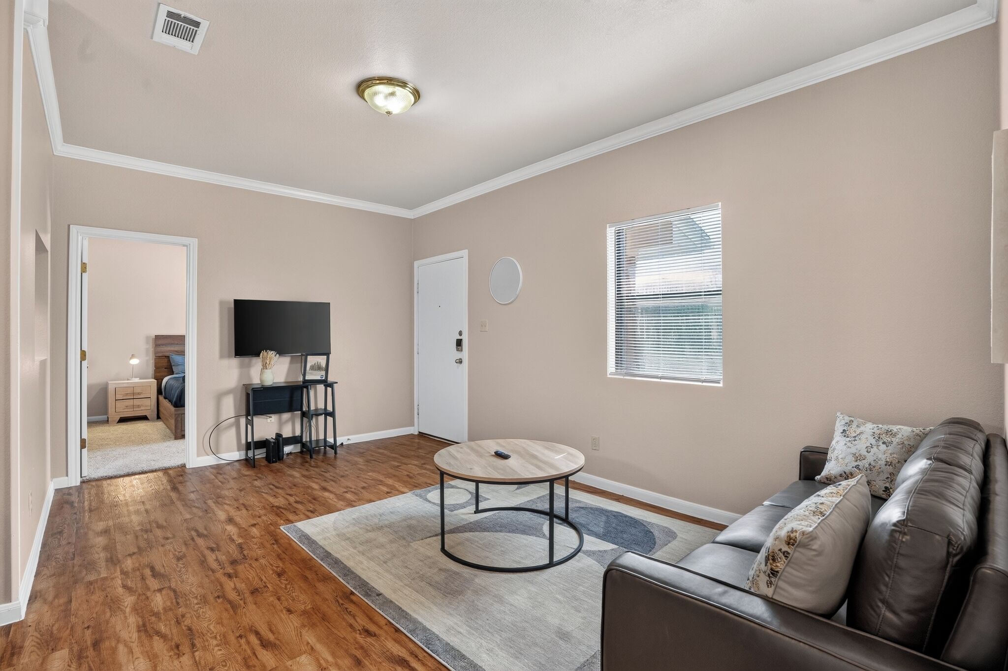 A living room with hardwood flooring, a round coffee table, a leather sofa, a television, and a window with blinds.