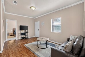 A living room with hardwood flooring, a round coffee table, a leather sofa, a television, and a window with blinds. - Bright & Comfortable Saddlebrook Apartment (Lumberton)