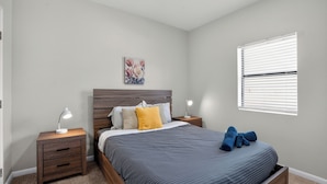 A bedroom with a bed, nightstands, a lamp, a painting, and a window with blinds. - Saddlebrook: Comfortable Furnished Apartment (Lumberton)