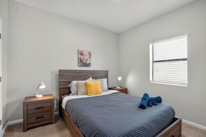 Ein Schlafzimmer mit einem Bett, Nachttischen, einer Lampe, einem Gemälde und einem Fenster mit Jalousien.
