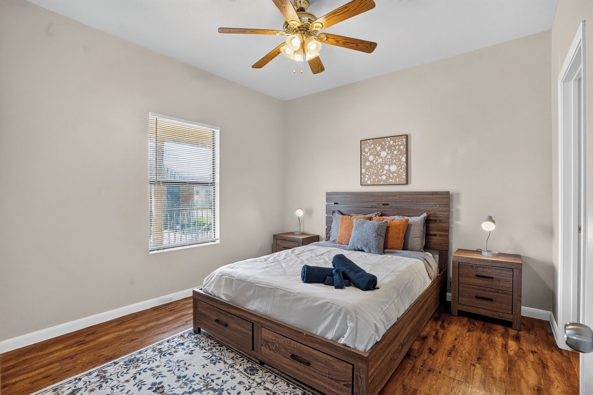 Une chambre à coucher avec un lit en bois, deux tables de chevet, un ventilateur de plafond, une fenêtre avec des stores et un tapis à motifs floraux.