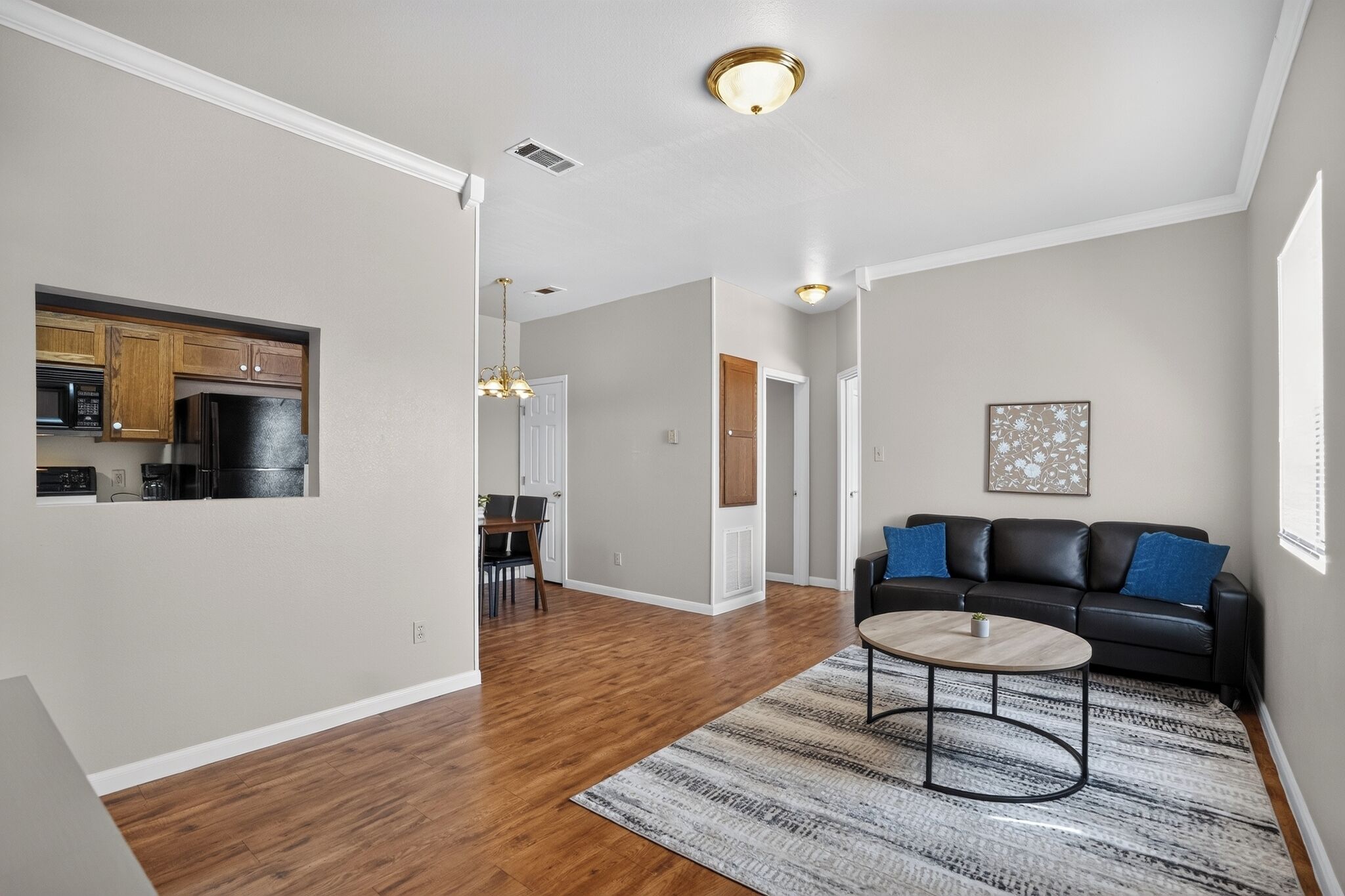 A modern apartment with hardwood flooring, a dining area, a kitchen with dark appliances, a living room with a black leather sofa, and a patterned rug.