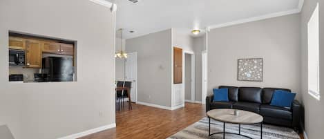 A modern apartment with hardwood flooring, a dining area, a kitchen with dark appliances, a living room with a black leather sofa, and a patterned rug.