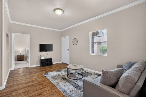 A living room with a flat-screen TV, a round coffee table, a sofa, and a window with blinds.