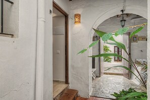 Un couloir étroit doté d’un sol carrelé, d’une porte en bois et d’un mur blanc orné d’un petit luminaire.