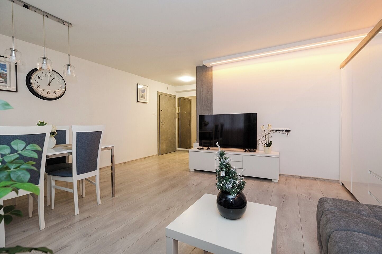 Un salon moderne avec un téléviseur à écran plat, un coin repas avec des chaises, un petit sapin de Noël et une horloge au mur.