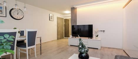 Una sala moderna con un televisor de pantalla plana, un comedor con sillas, un pequeño árbol de Navidad y un reloj en la pared.