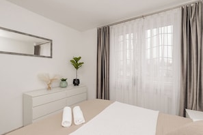 Ein modernes Hotelzimmer mit Bett, Nachttisch mit Dekoration, großem Fenster mit Vorhängen und Spiegel an der Wand.