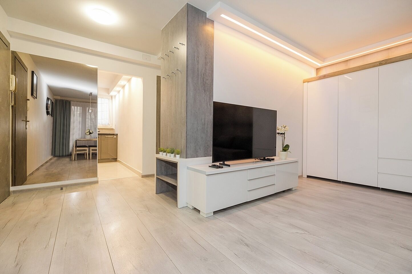 A modern living room with a flat-screen TV, a white entertainment unit, and a dining area in the background.