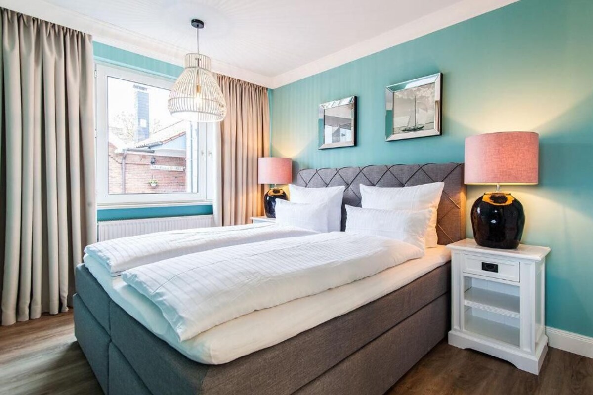 A bedroom with a large bed, two bedside tables, a window with curtains, and wall-mounted lamps.