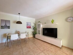 Una sala de estar con televisor de pantalla plana, una mesa de comedor con sillas, un armario blanco y una decoración de cactus.