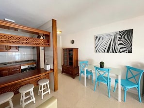 Eine Küche mit Holzbalken, weißen Barhocker, einem Esstisch mit blauen Stühlen und einem Wandbild mit Zebra-Muster.