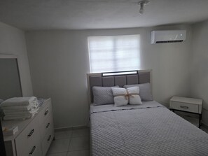 A bedroom with a bed, a nightstand, a dresser, a window with blinds, and an air conditioning unit.