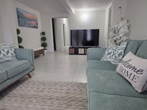 A modern living room with a grey sofa, a TV, and a 'Welcome Home' pillow. - Perfect 4-bedroom house with AC in charming Humacao (Humacao)