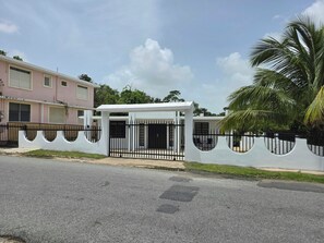 A gated residential area with a white fence, a covered entrance, and a pink building in the background.