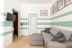 Ein Zimmer mit einer gestreiften Wand, einem grauen Sofa mit Kissen, einem am Wand montierten Flachbildfernseher und einer Tür mit grünem Rahmen.