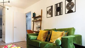 Un divano verde con cuscini decorati, un tavolino in legno con della frutta e opere d'arte astratte alle pareti.