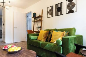 Ein grünes Sofa mit gemusterten Kissen, ein Holztisch mit Obst und abstrakte Wandbilder.
