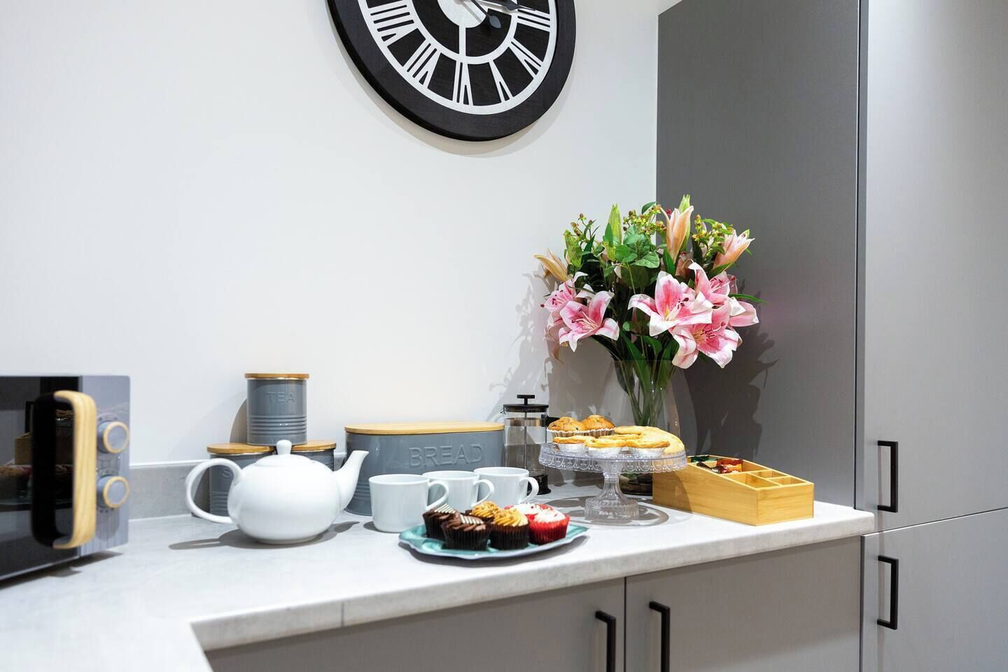 Una encimera de cocina con un reloj, juego de té, tazas, un florero con flores y una bandeja de pasteles.