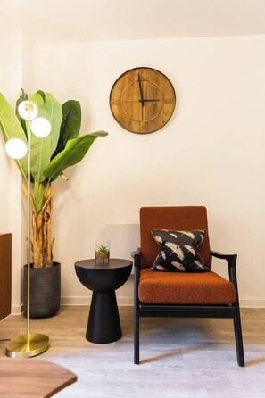 A room with a wooden wall clock, a brown armchair with a patterned cushion, a tall plant with large leaves, a small black side table with a glass top, and a floor lamp with three white globes.