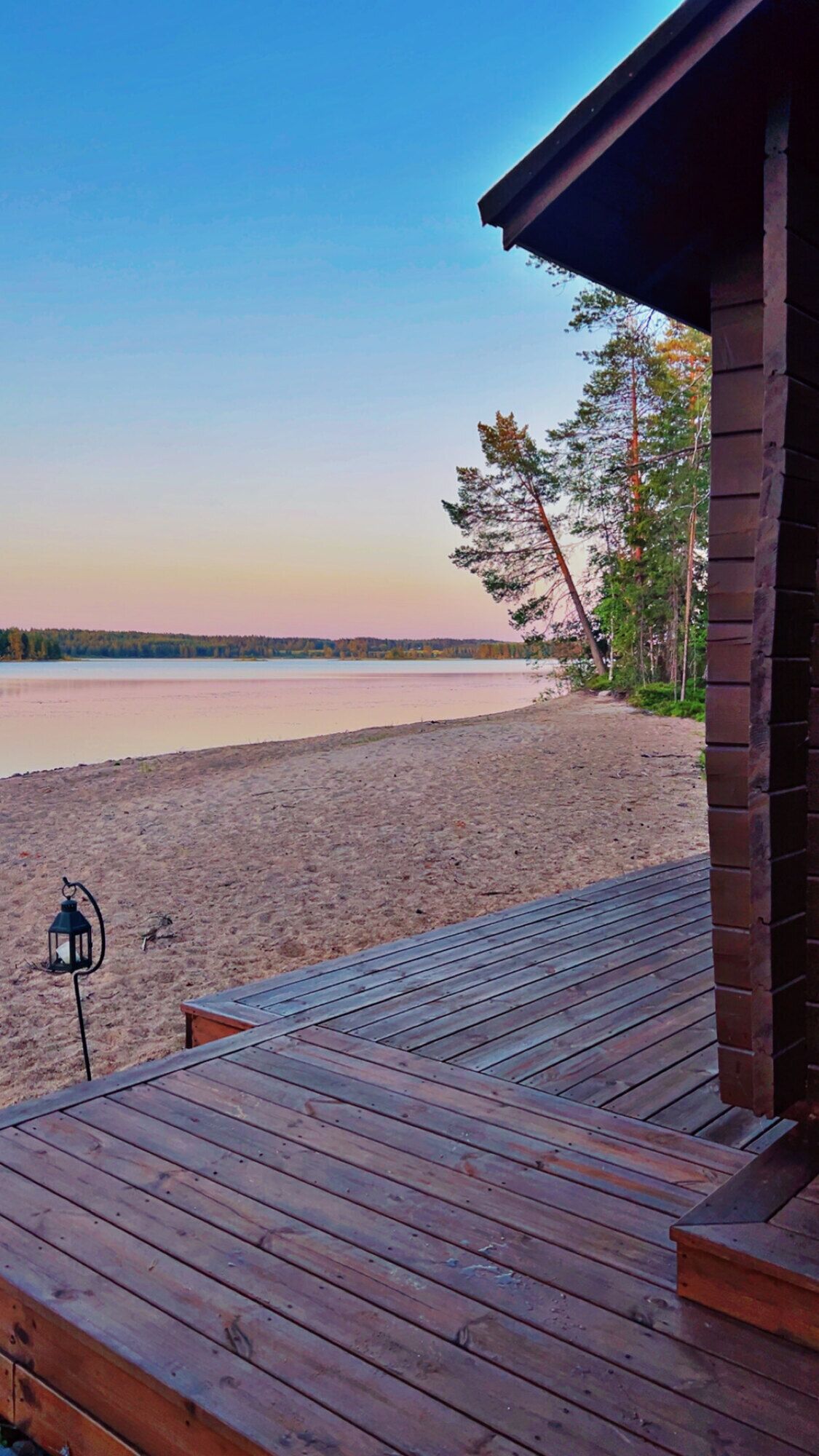 Una terraza de madera que da a una playa, con una vista despejada de un lago y árboles al fondo.