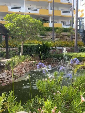 Ein angelegter Garten mit Teich, verschiedenen Pflanzen und einem Gebäude im Hintergrund.