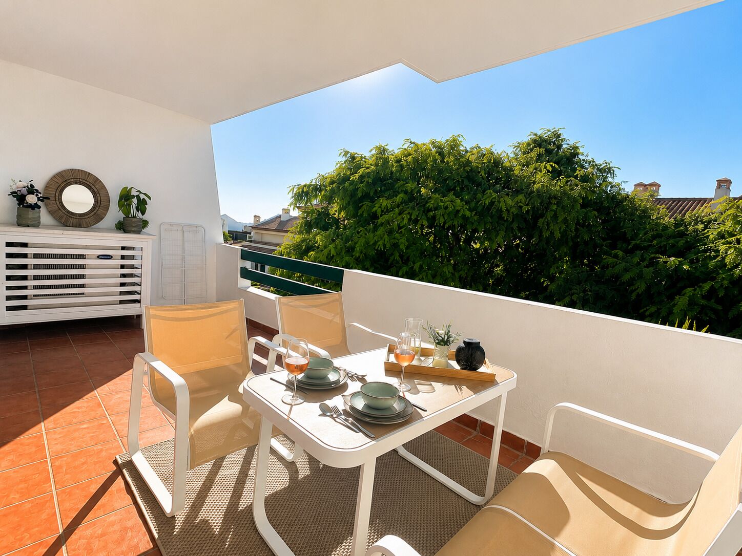 Un balcón con una mesa lista para comer, sillas, un radiador blanco y vistas a zonas verdes y edificios.