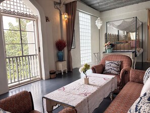 Ein Zimmer mit einem großen Fenster, einer Couch, einem Tisch mit Tischdecke und einem Bett mit gemusterten Betttextilien.