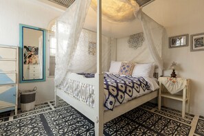 Ein Schlafzimmer mit einem Himmelbett, einem Nachttisch mit Lampe, einem Spiegel und einem an der Wand hängenden Kunstwerk.