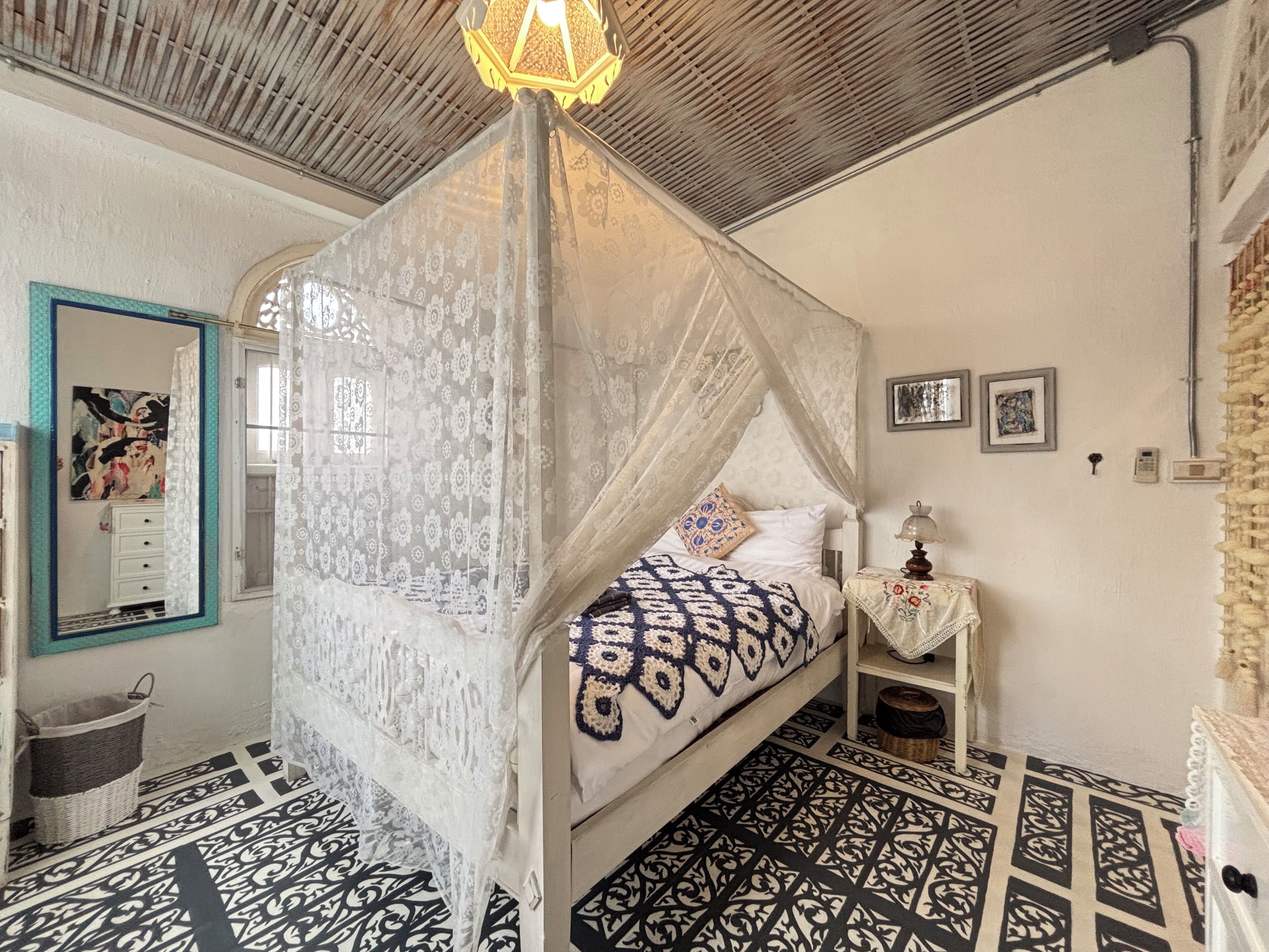 Een kamer met een bed onder een hemelbed, een nachtkastje met een lamp, een telefoon aan de muur en een vloer met een patroon.