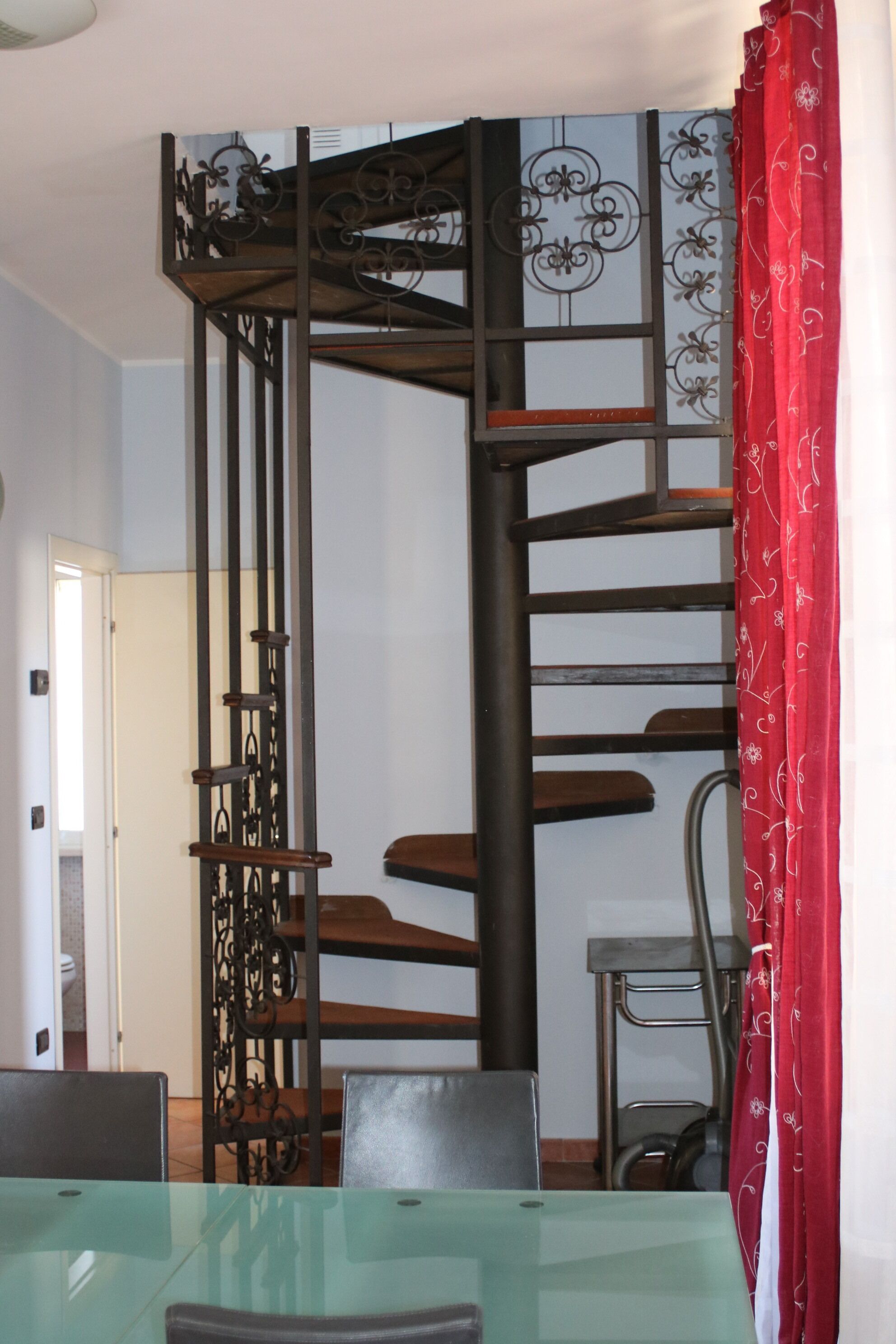 A dining area with a glass table, metal spiral staircase, and red curtains.