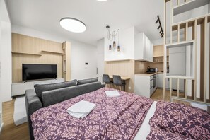 A modern hotel room with a large bed, a flat-screen TV, a desk, a chair, a kitchenette, and a wardrobe.
