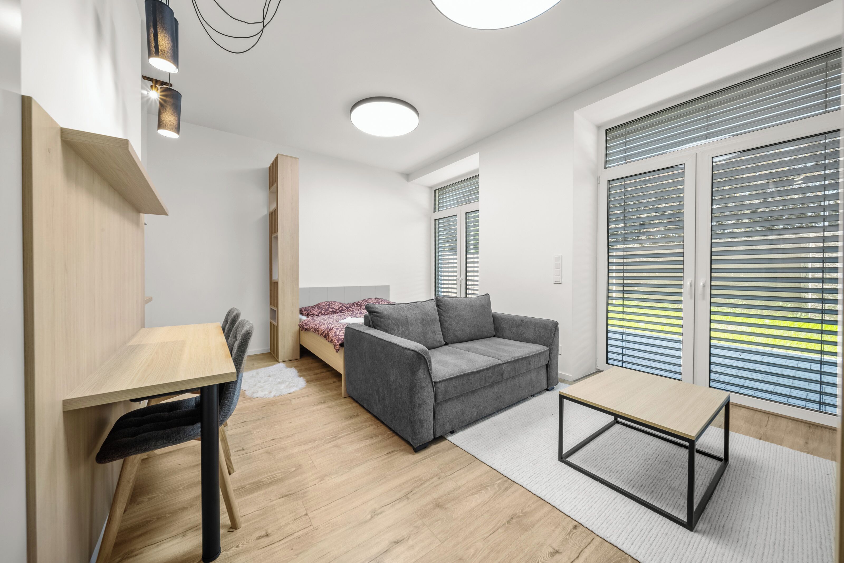 A modern, well-lit room with a bed, a grey sofa, a small table, and a desk.