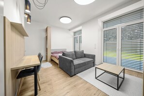 Ein modernes, helles Zimmer mit einem Bett, einem grauen Sofa, einem kleinen Tisch und einem Schreibtisch.