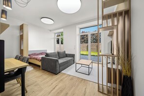 A modern living room with a sofa, a coffee table, and a TV. There is a dining area with a table and chairs. The room has large windows and a view of the outdoors. - 1 room apartment with garden, Nálepkova 1390 (Liptovský Mikuláš)