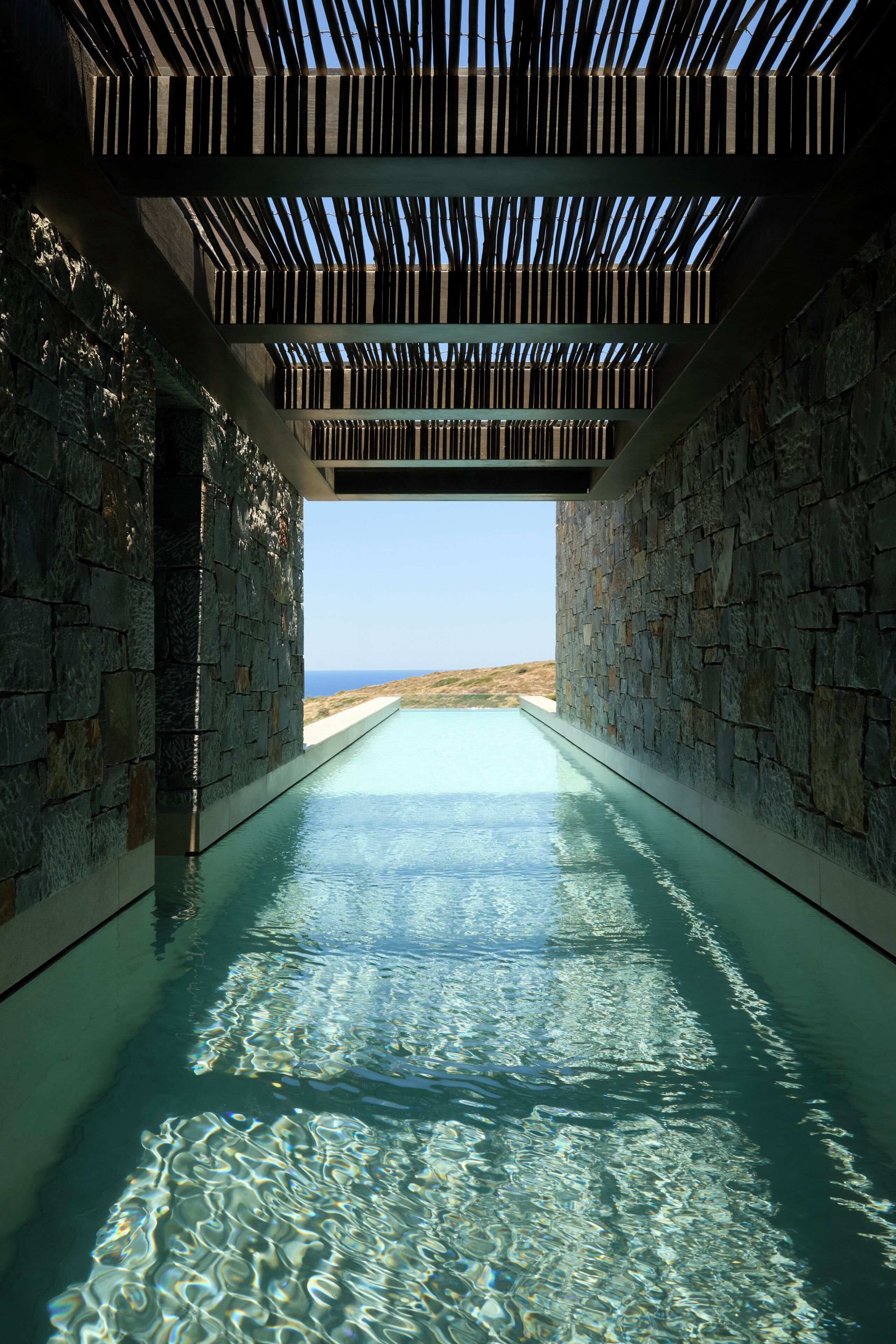 An infinity pool with a clear view of the ocean, surrounded by stone walls and a wooden ceiling structure.
