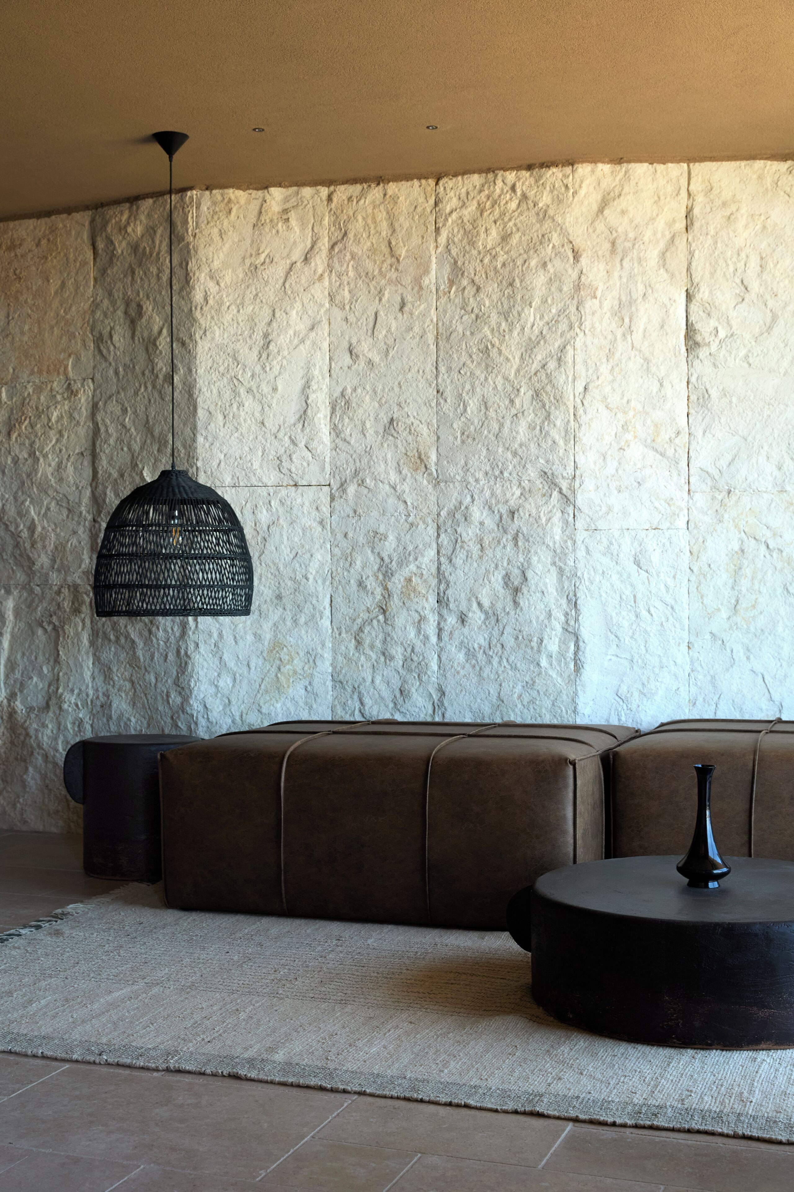 A modern interior with a stone wall, a woven pendant light, a leather sofa, and a round table.
